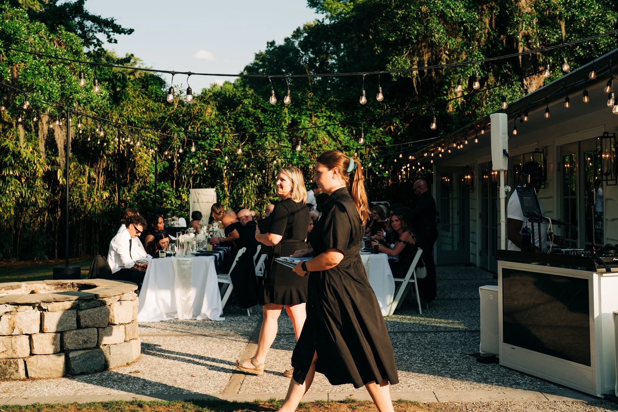 Outdoor event with people sitting at tables, waitstaff serving food, string lights hung overhead, and a DJ setup nearby, surrounded by lush green trees.