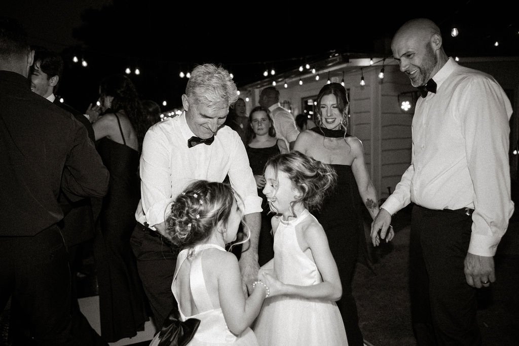 People dancing and celebrating at a formal event or wedding reception, including two young girls in white dresses and several adults in formal attire.