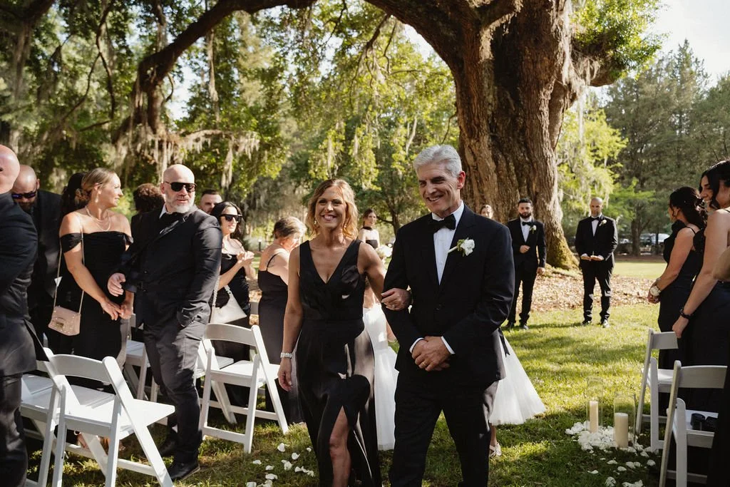 A man in a tuxedo walking down the aisle at an outdoor wedding ceremony, smiling, with a woman in a black dress supporting him, surrounded by seated guests in black attire under a large tree.
