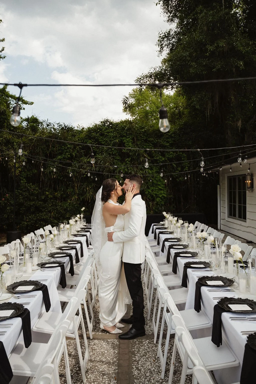 A bride and groom sharing a kiss at an outdoor wedding reception with long tables decorated with black and white table settings and string lights overhead.
