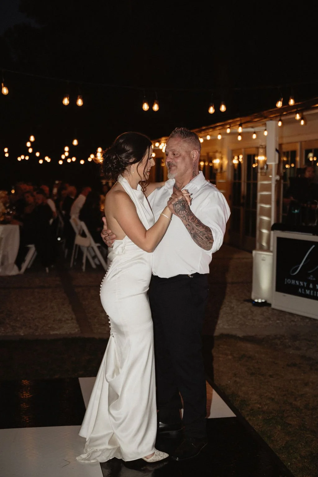 A bride and groom dancing at their wedding reception during the evening, under warm string lights outside a venue with guest tables in the background.