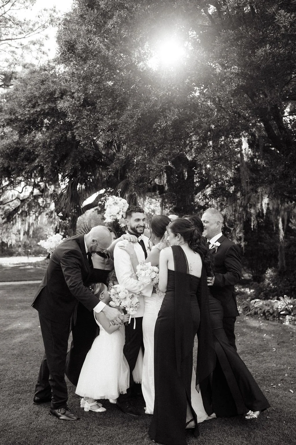 Black and white photograph of a wedding party outdoors under a large tree with sunlight filtering through. The group includes adults and children, with the bride and groom in the center, surrounded by bridesmaids and groomsmen, all smiling and celebr