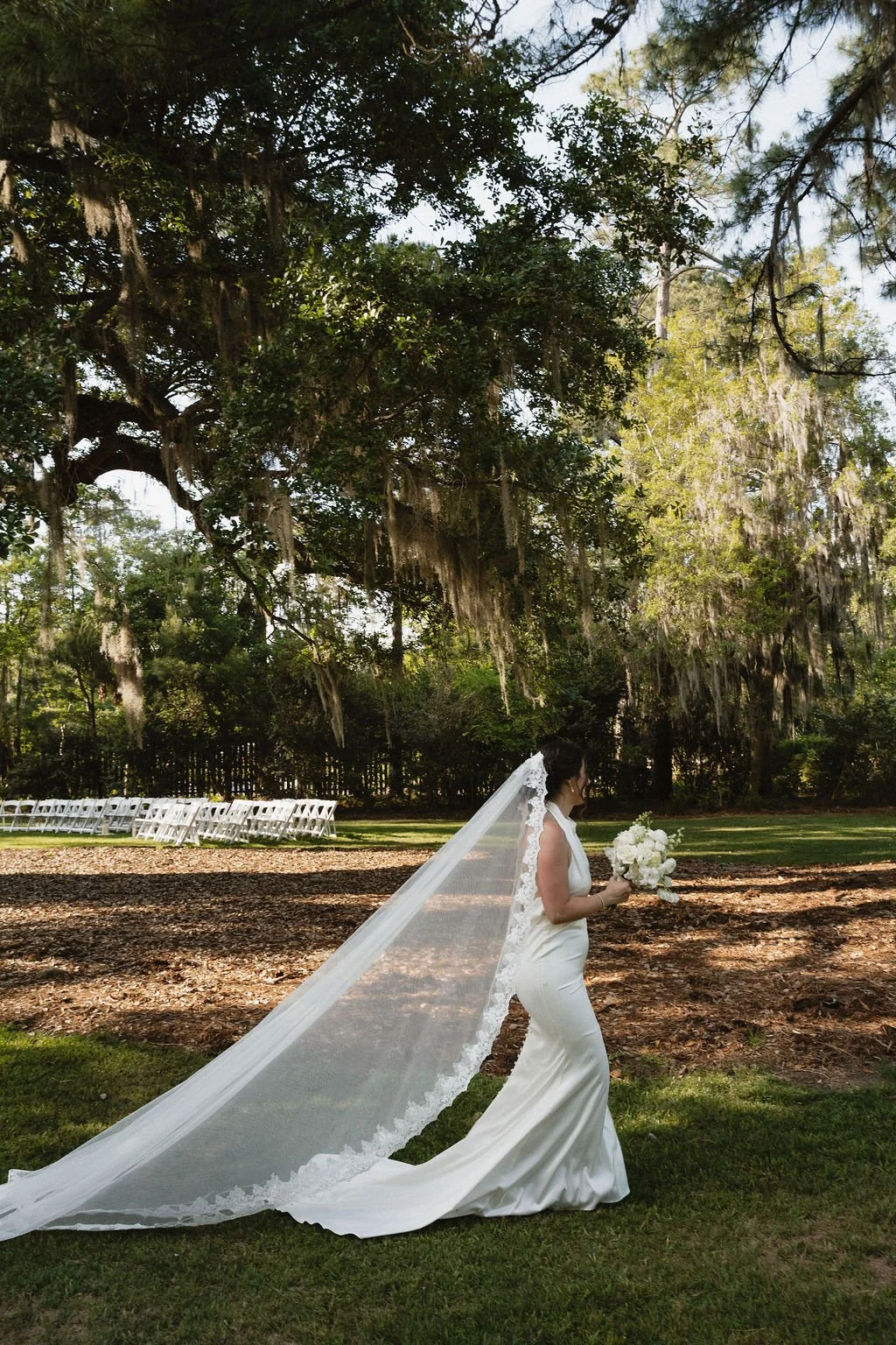 A bride in a white wedding gown and veil holding a bouquet of white flowers standing outdoors beneath large trees with hanging moss, with empty white chairs arranged in rows in the background.