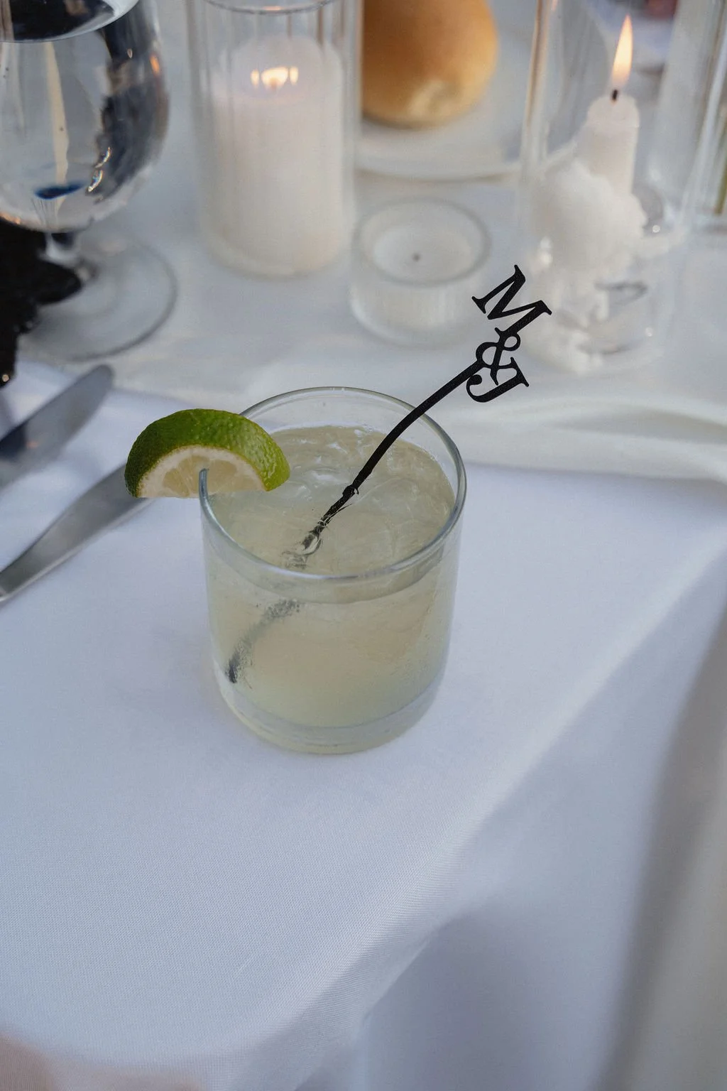 A glass of lemonade with a lime wedge on the rim and a decorative stir stick with 'M & J' on it, on a table with a white tablecloth and candles in the background.