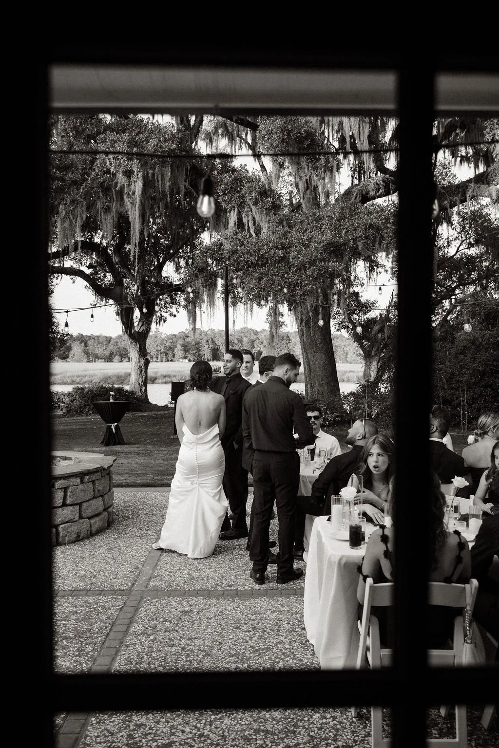 Black and white photo of a wedding reception outdoors, viewed through a window. Bride in a strapless wedding gown is talking to a man in dark attire. Several guests seated at tables are visible, with some engaged in conversations. Large trees and han