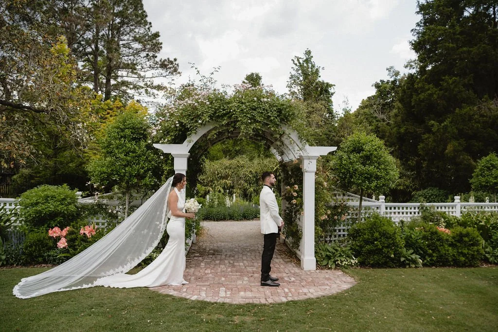 Bride and groom standing apart on wedding day, outdoor garden setting with archway and lush greenery.
