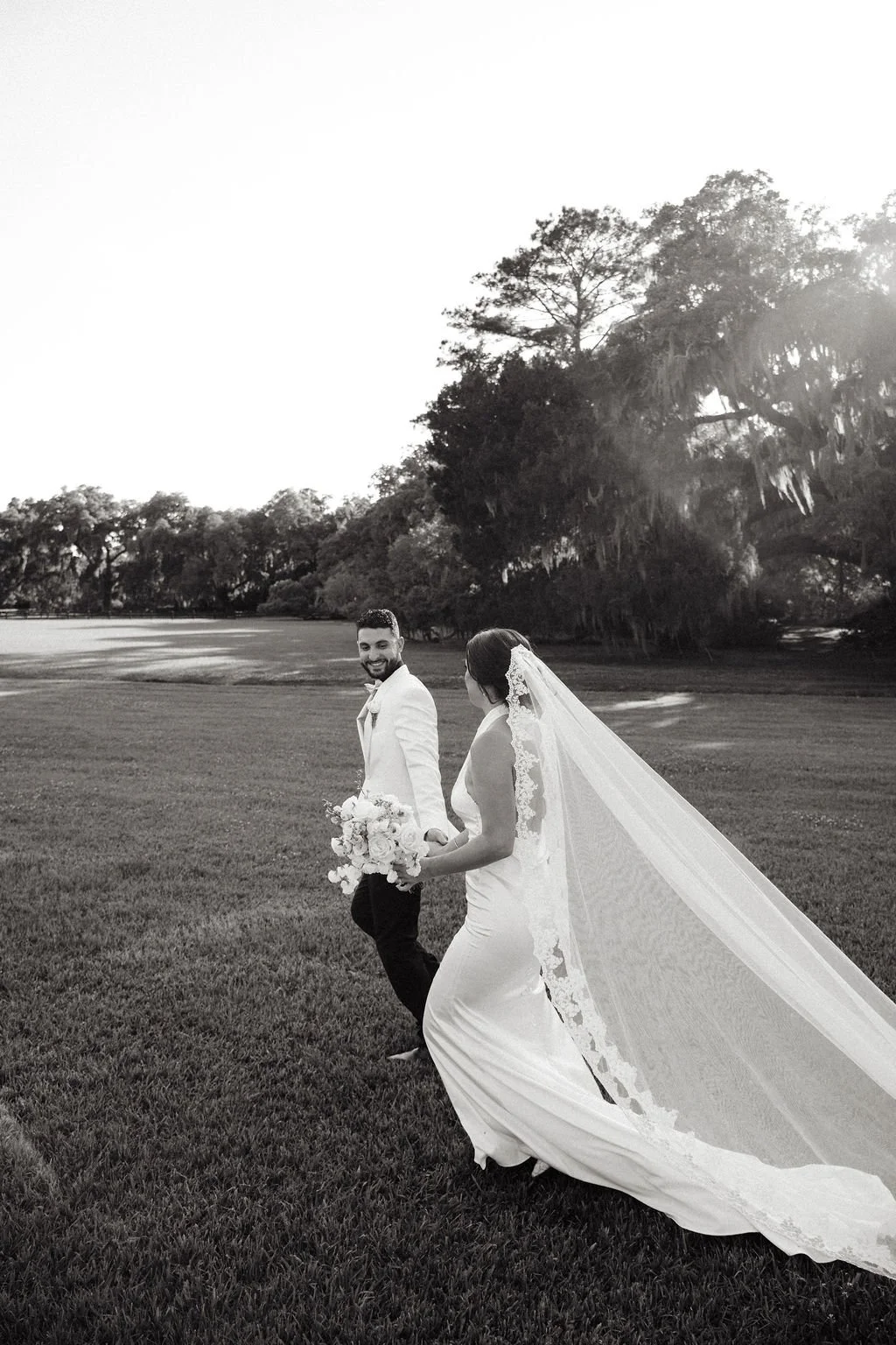 Black and white photo of a smiling bride and groom walking together in a park, with the bride wearing a wedding dress and veil and holding a bouquet, and the groom in a suit, under large trees with Spanish moss.