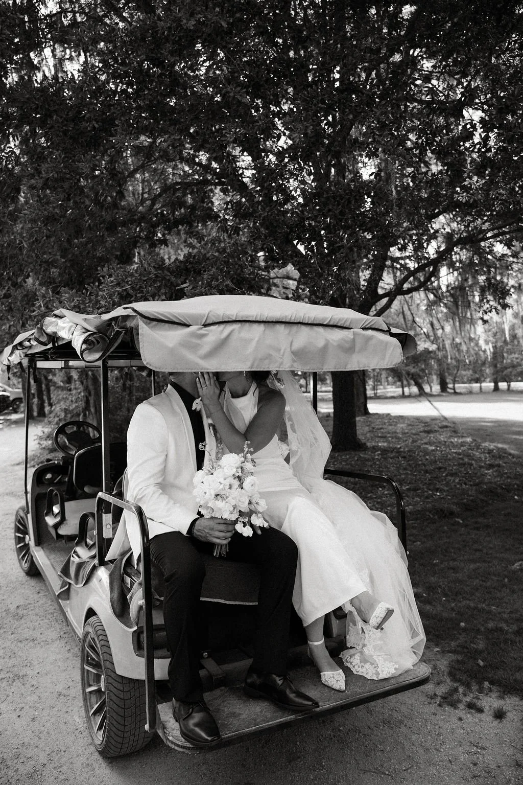 A bride and groom sitting close together on a golf cart, kissing under a canopy with trees in the background. The groom holds a bouquet of flowers. The bride is wearing a wedding gown and heels, the groom is in a suit.