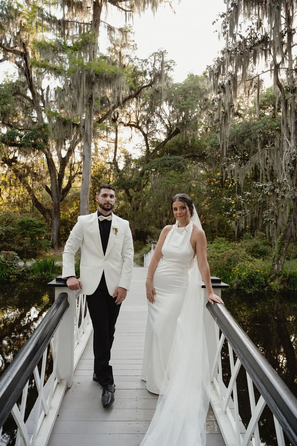 A bride and groom standing on a white bridge over a pond, surrounded by trees with Spanish moss, during sunset or early evening.