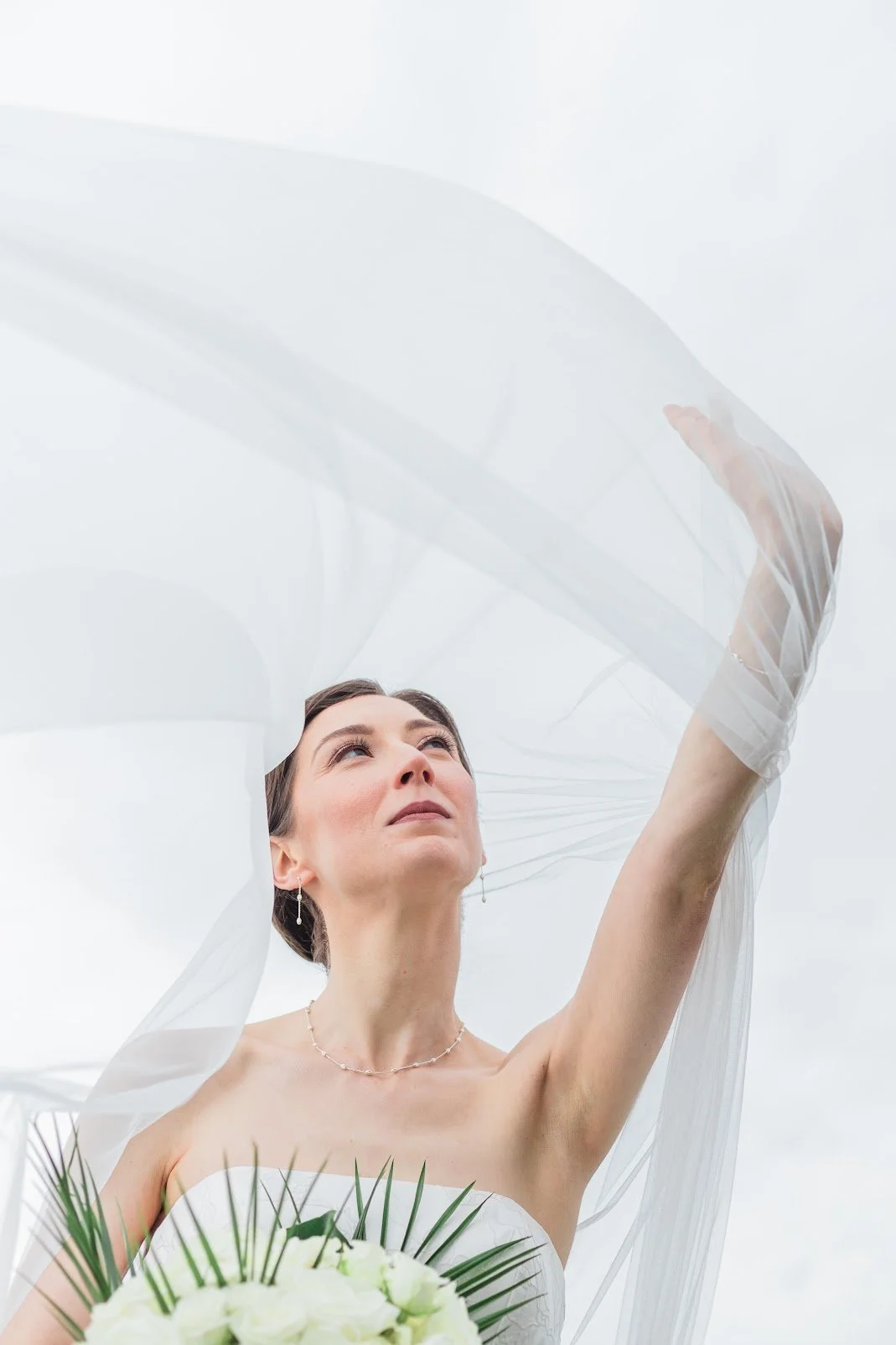Bride looking upward with veil flowing around her, holding a bouquet of white flowers and greenery on a cloudy day.