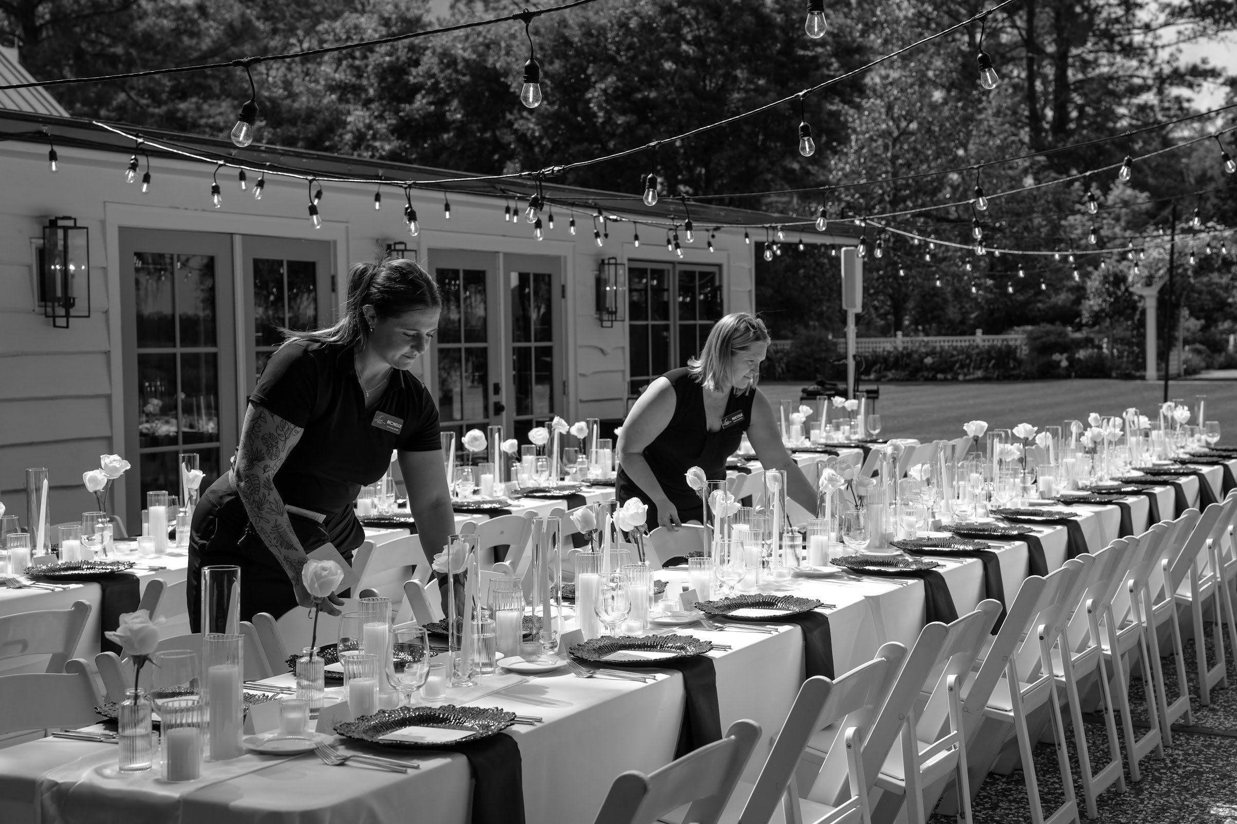 Two women setting up a long outdoor banquet table with white tablecloths, candles, and floral centerpieces, under string lights, in front of a house with large windows and a lawn.