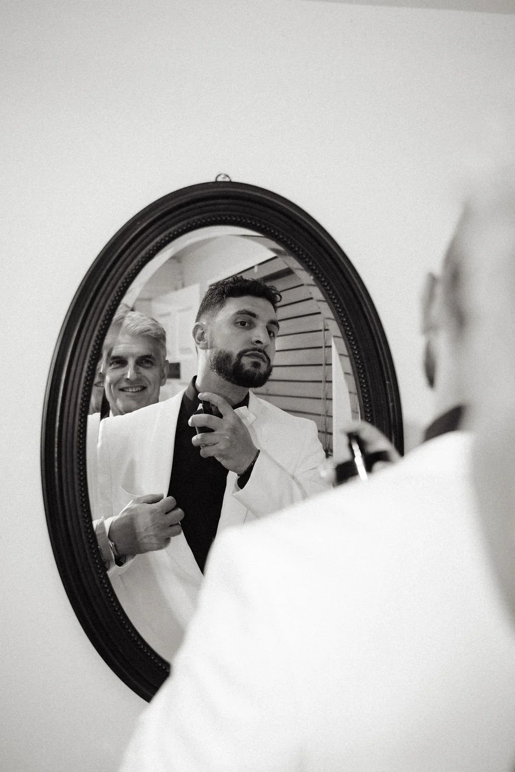 A man in a white suit taking a selfie in a mirror, with another man smiling behind him in the background.