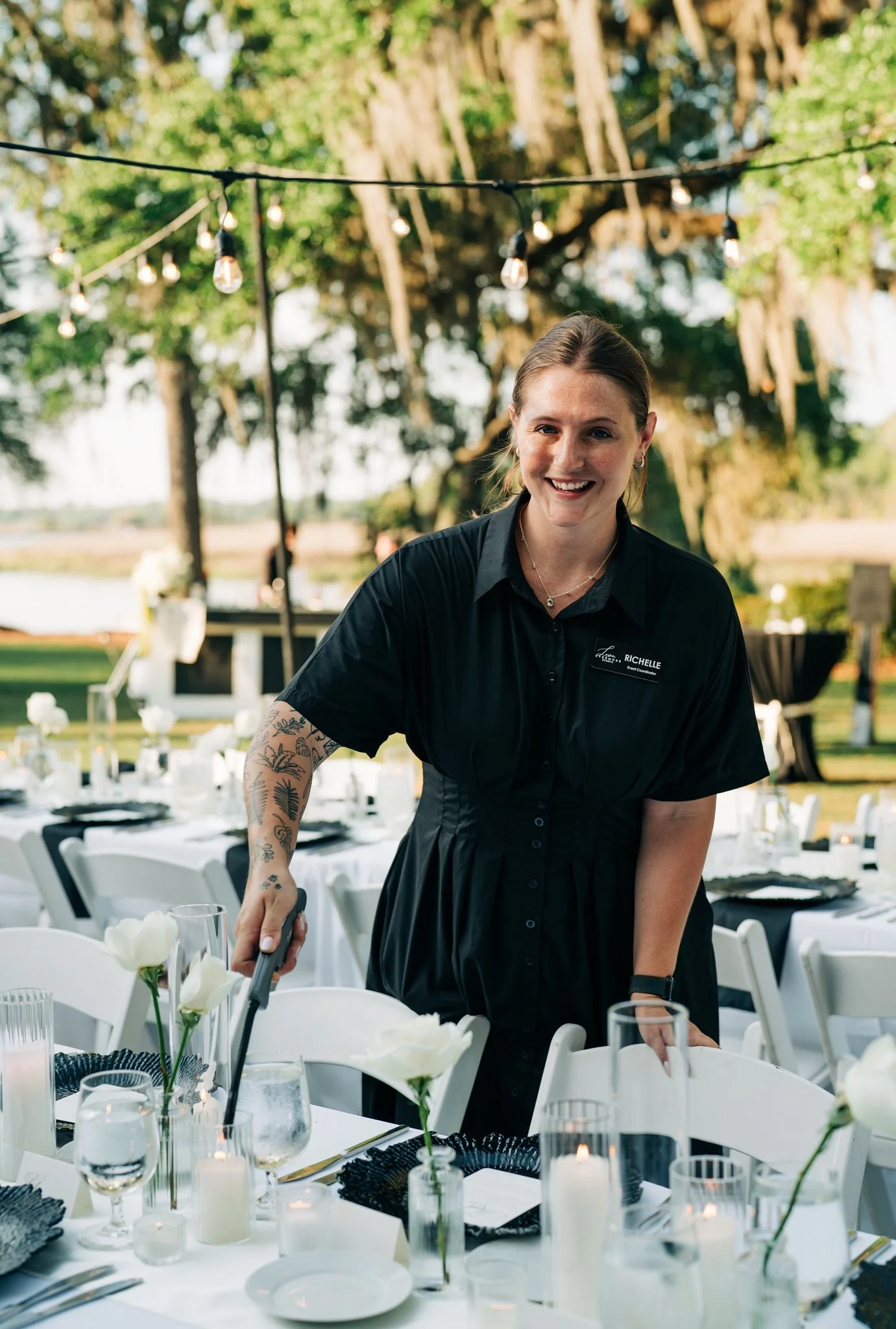 A smiling woman with a tattooed arm and a name tag that reads 'RICHELLE' is setting a table outdoors during daylight. She is dressed in a black uniform and is surrounded by decorated tables with white tableware, flowers, and candles, under string lights and large trees.