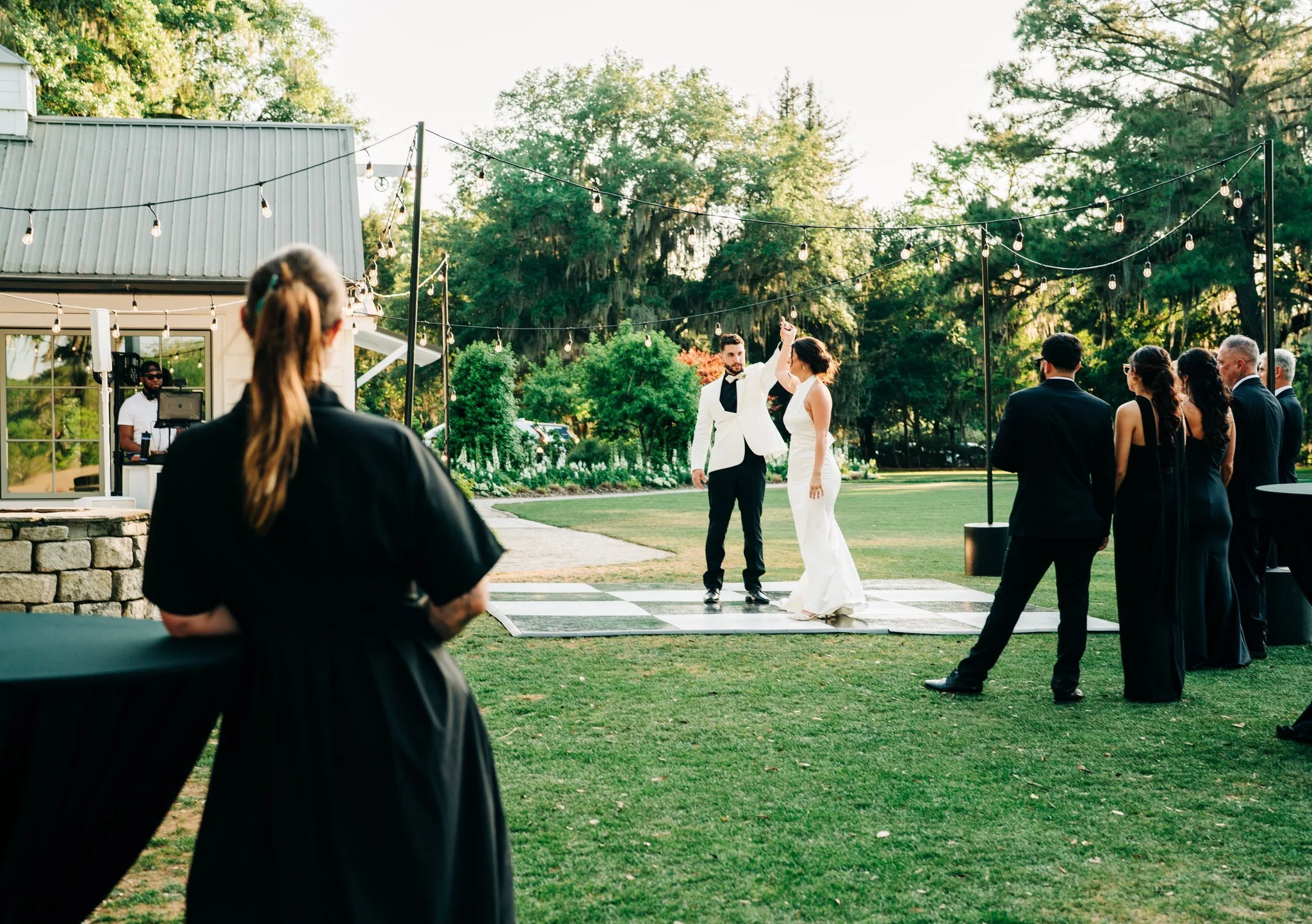 A bride and groom dancing outdoors during a wedding ceremony with guests watching, string lights hanging above, in a lush garden setting.