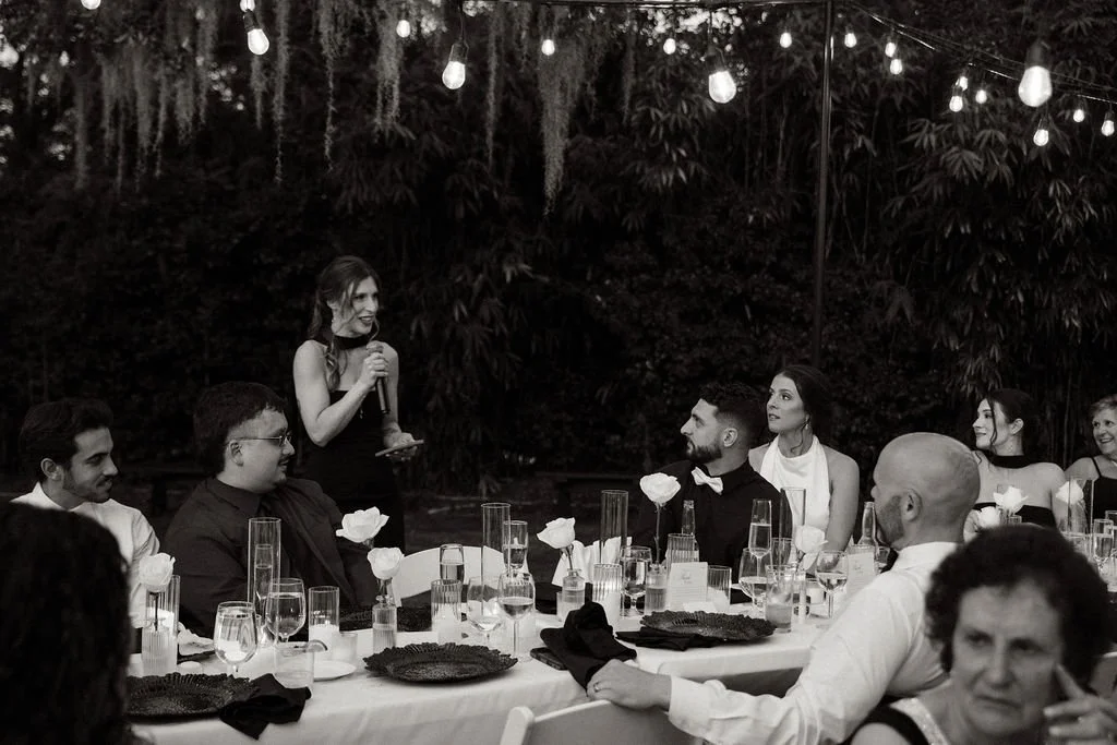 A woman giving a speech or toast at an outdoor evening event, with guests seated at a table decorated with flowers and glassware, under string lights.