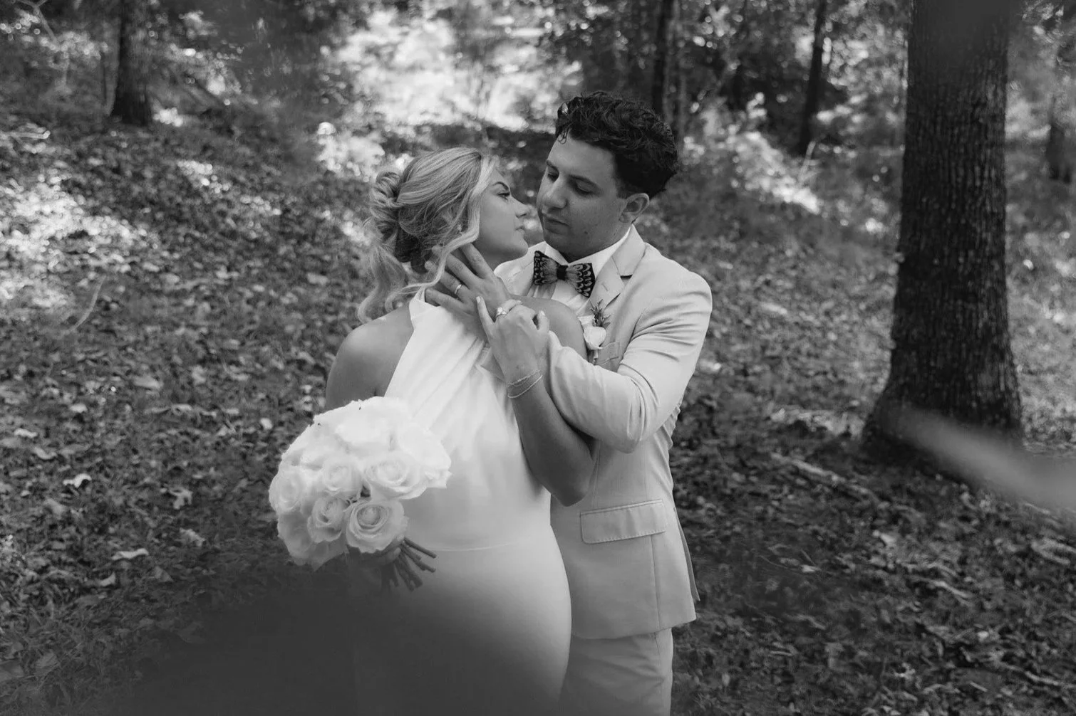 A black-and-white photo of a couple in wedding attire in a wooded area. The woman is holding a bouquet of roses, and the man is holding her face as they gaze into each other.