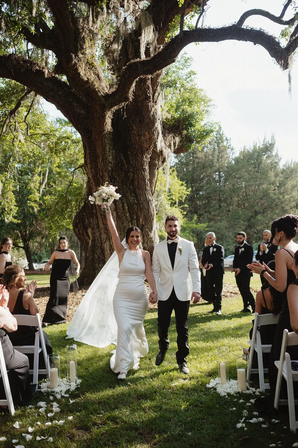 Bride and groom walking down the aisle outdoors during their wedding ceremony, surrounded by guests clapping and celebrating under a large tree.