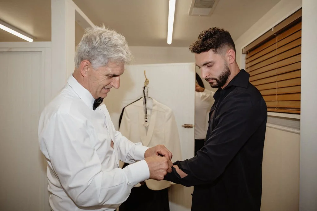 An older man in a white dress shirt and black bow tie helping a younger man in a black shirt with his sleeve cuff in a dressing room or backstage area.