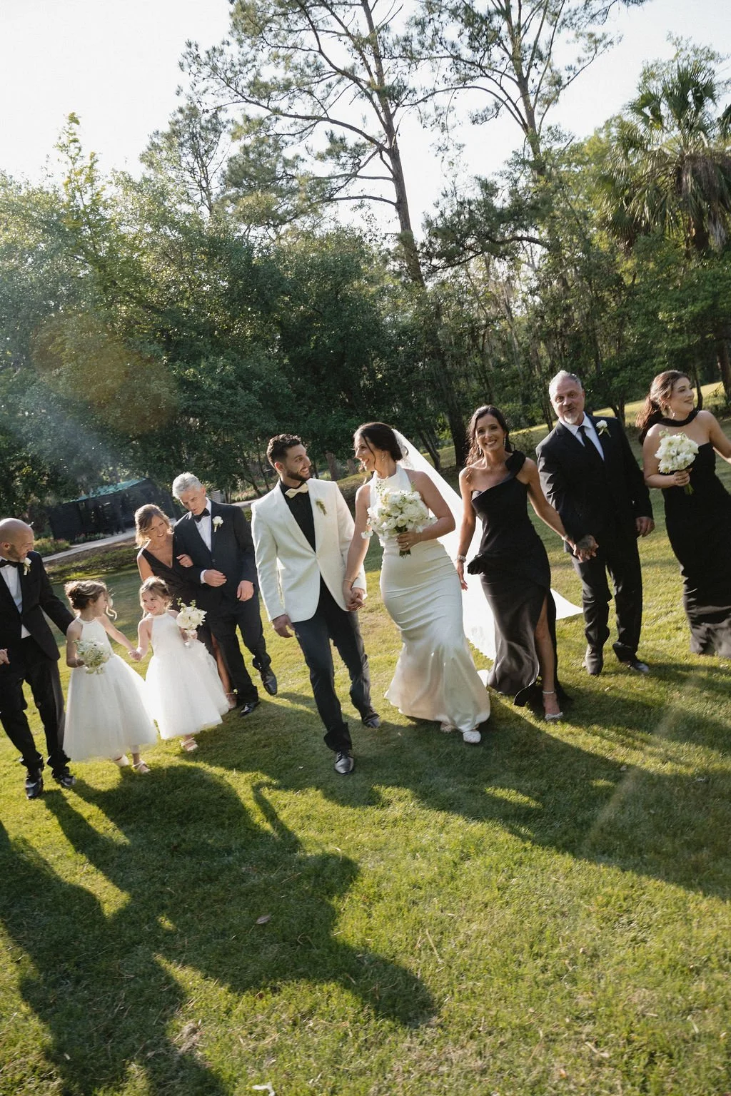 A wedding party walking outdoors on grass, consisting of bride, groom, children, and guests, with trees and sunlight in the background.