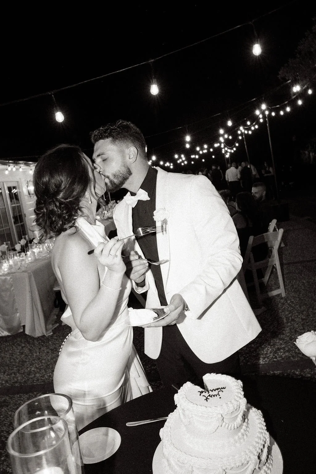 A black and white photo of a newlywed couple sharing a kiss at their wedding reception, surrounded by string lights and guests, with a wedding cake visible in the foreground.