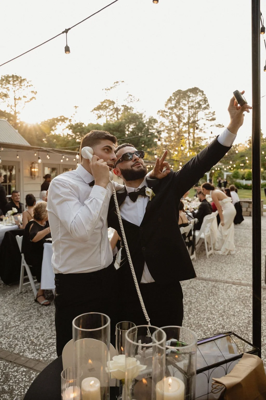 Two men, one in a tuxedo and sunglasses and the other in a white shirt, are taking a selfie with a pole while at a wedding reception outdoors during sunset. The scene is decorated with string lights and tables filled with guests.