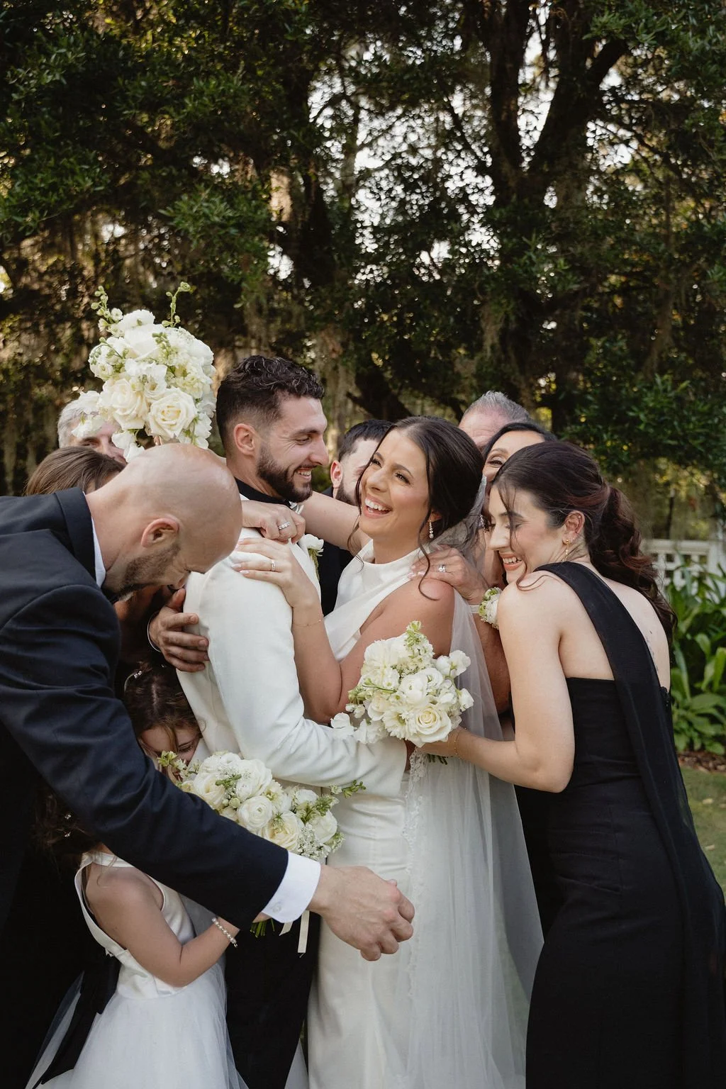 Group of people, including bride and groom, celebrating at a wedding outdoors with trees in the background, all smiling and hugging.