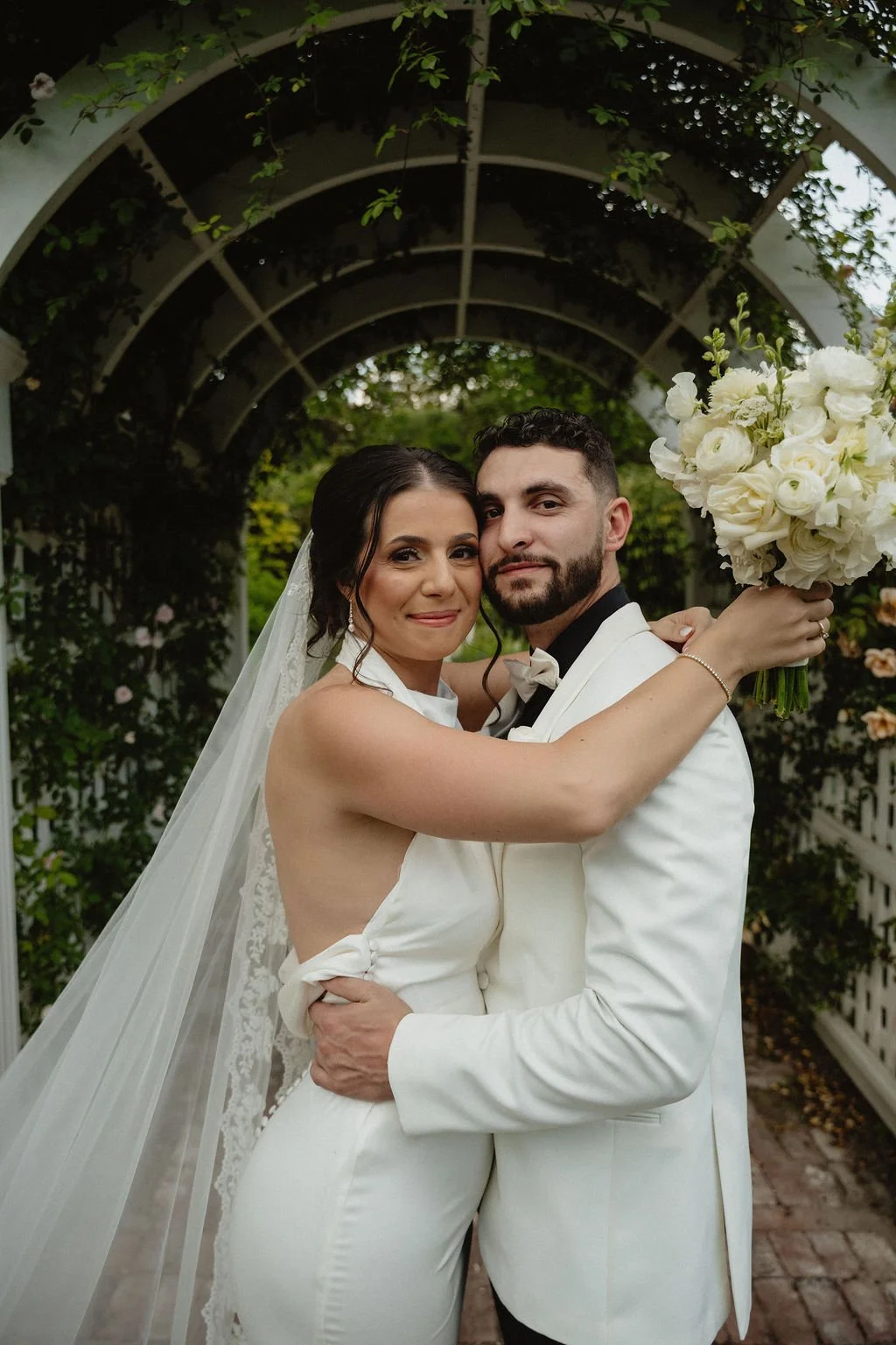 A newlywed couple in wedding attire embrace under a garden archway, with the bride holding a bouquet of white flowers.