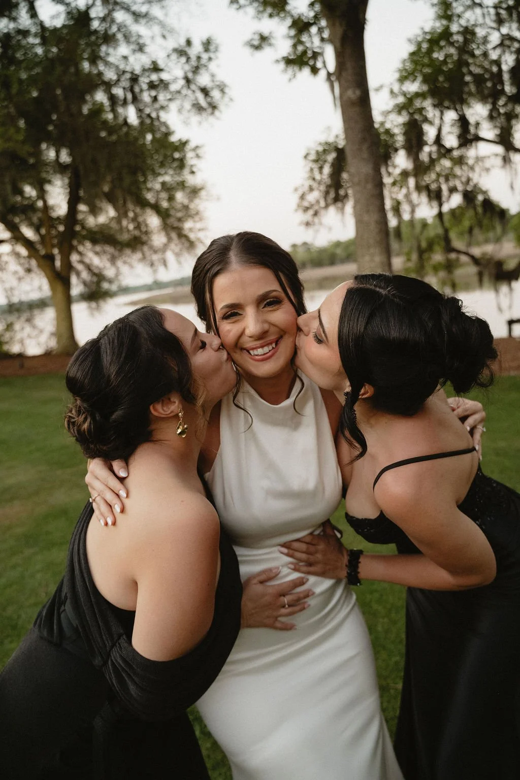 A woman wearing a white dress being kissed on each cheek by two women in black dresses outdoors near a lake and trees