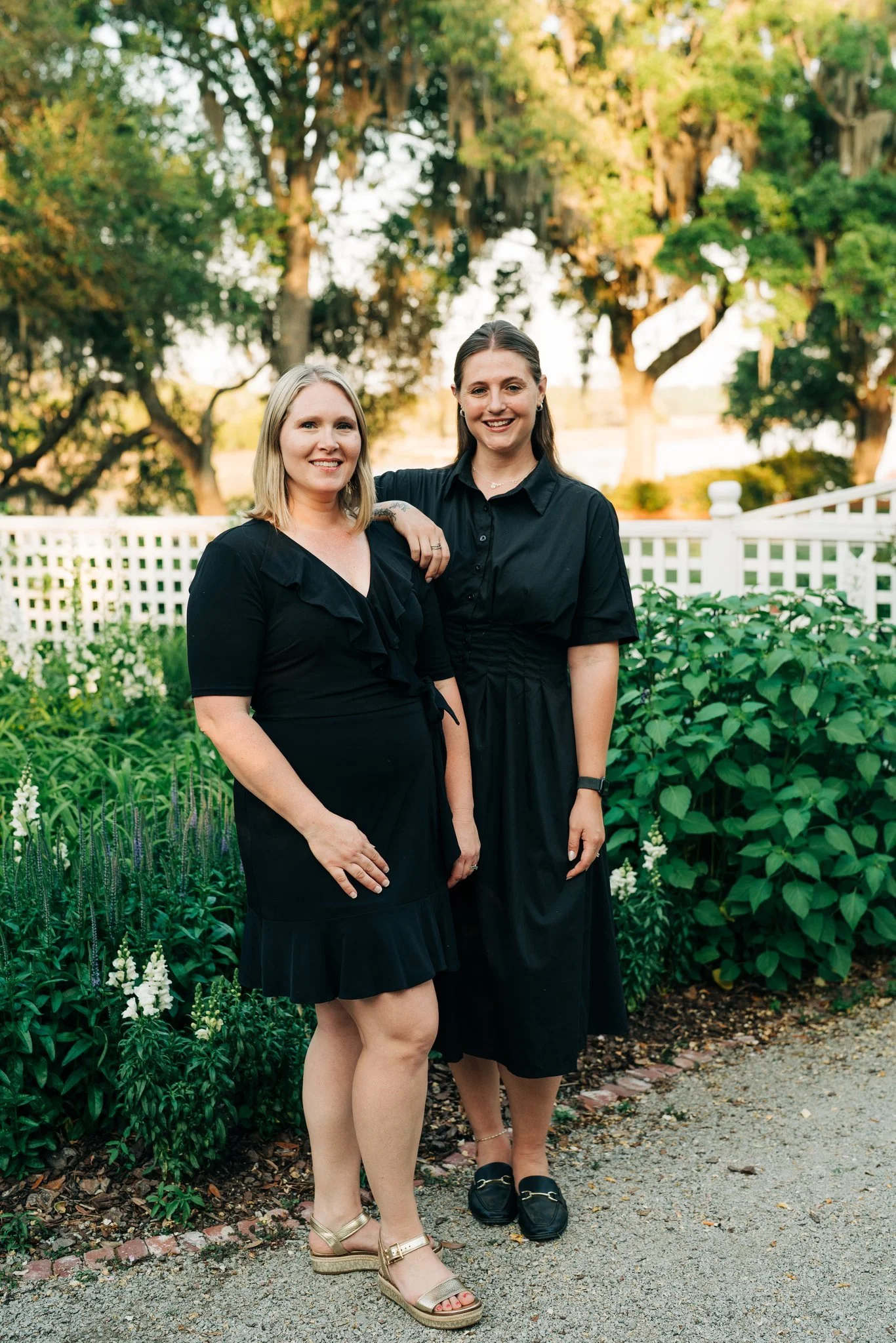Two women standing outdoors in a garden with green bushes and trees, wearing black dresses, smiling at the camera.