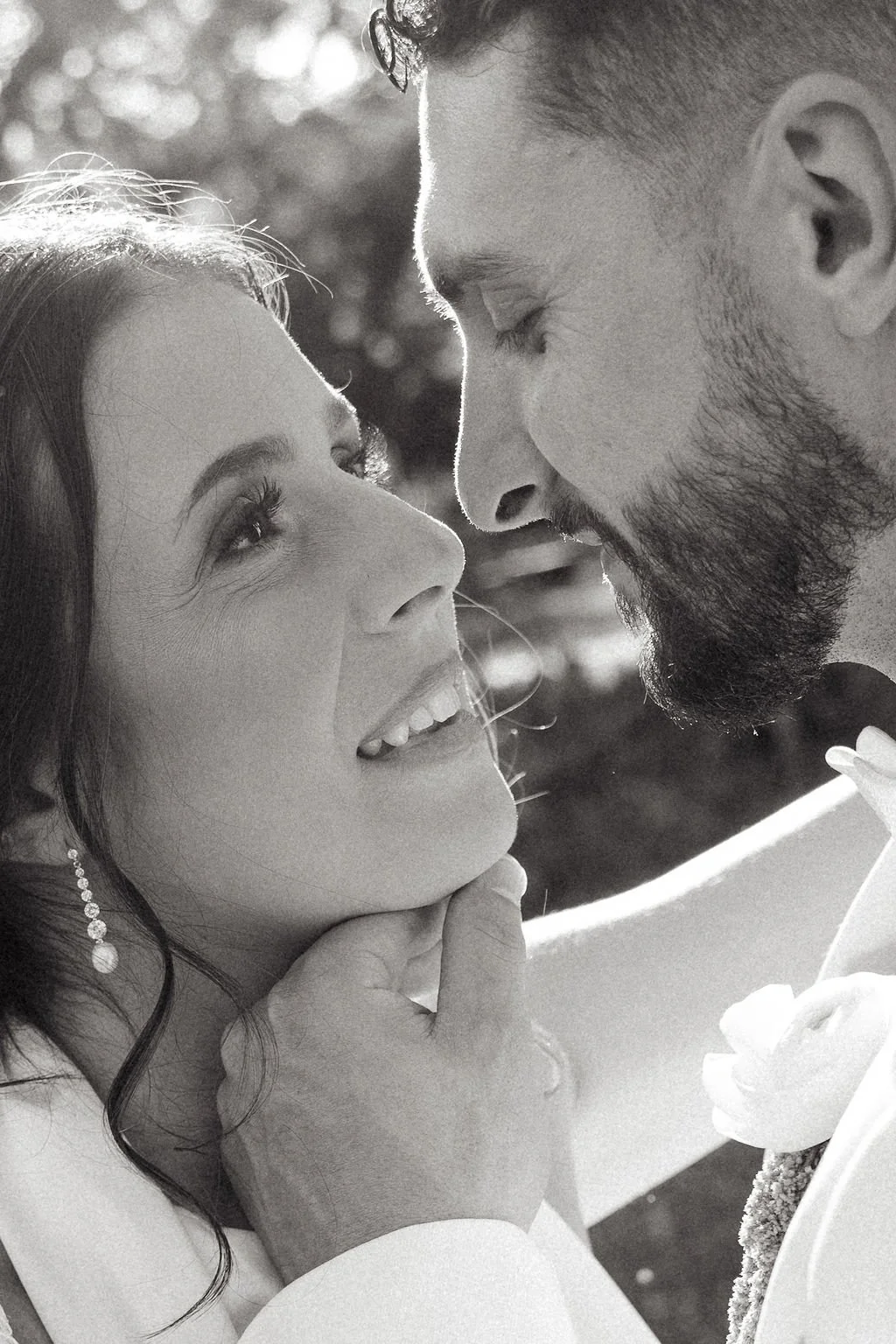 A black and white close-up photograph of a couple looking into each other's eyes, with their faces close together. The woman is smiling softly, and the man's face is partly shadowed with a beard. The woman's hand gently touches her chin, and she is w