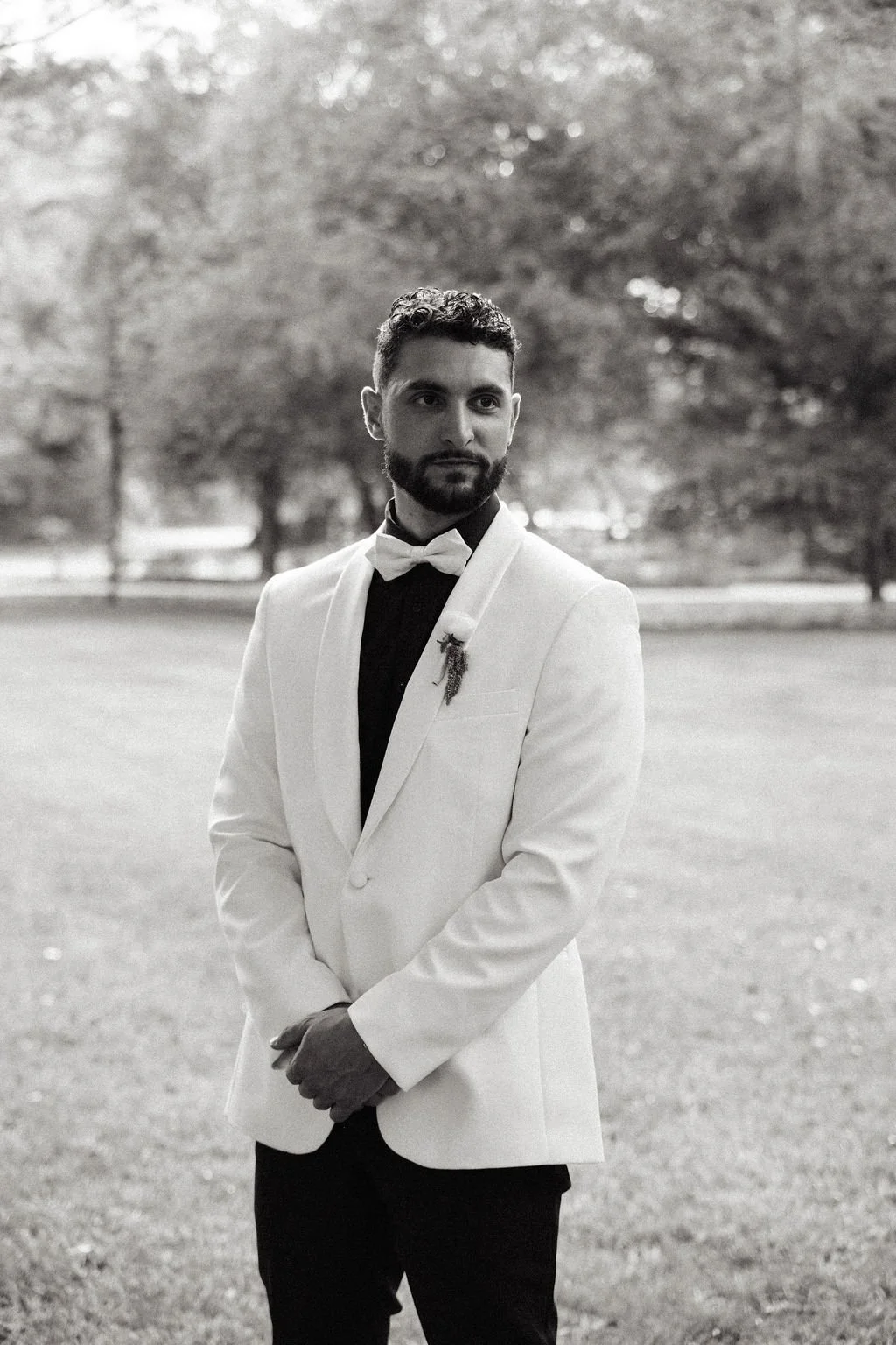 A man in a tuxedo standing outdoors, wearing a white jacket, black shirt, and bow tie, with a boutonniere on his lapel, in a park with trees in the background.
