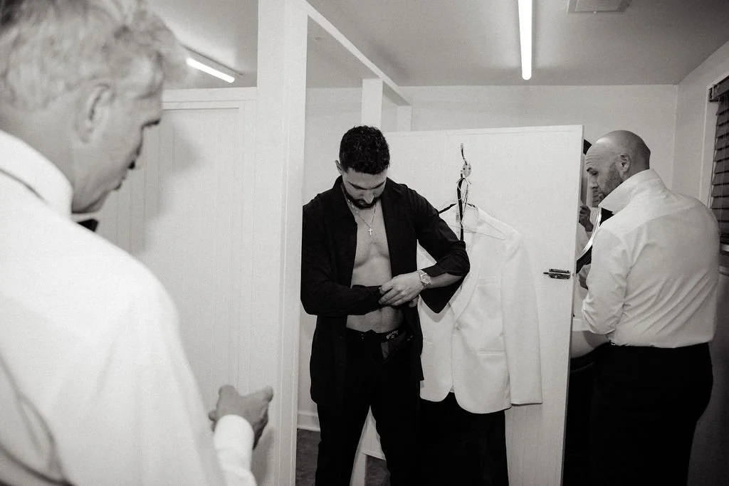 A man with sunglasses and an open shirt revealing a cross necklace is adjusting his shirt in front of a mirror. Two other men in white shirts are observing him inside a small room, possibly a dressing area or behind-the-scenes space.