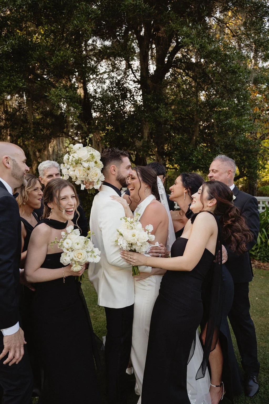Bride and groom smiling with wedding party in an outdoor garden setting surrounded by trees.