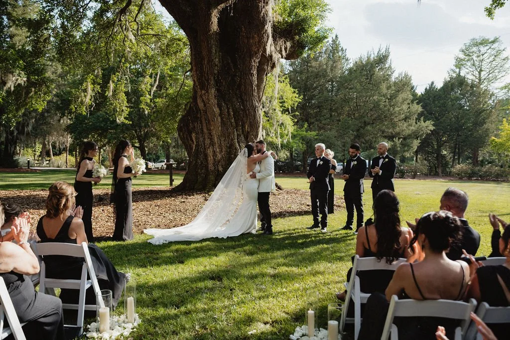 A wedding ceremony outdoors under a large tree with the bride and groom kissing, surrounded by bridesmaids, groomsmen, and seated guests in the afternoon sunlight.