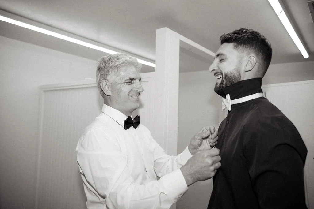 An older man adjusting a young man's tuxedo collar in a well-lit room, both smiling.