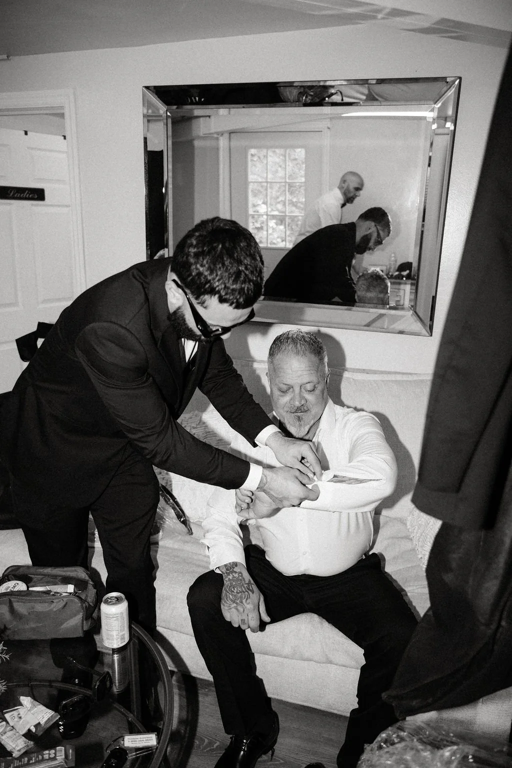 A man getting his shirt cuff adjusted by another man in a suit, while sitting on a couch. Reflections in a large mirror behind show more people in a room with natural light.