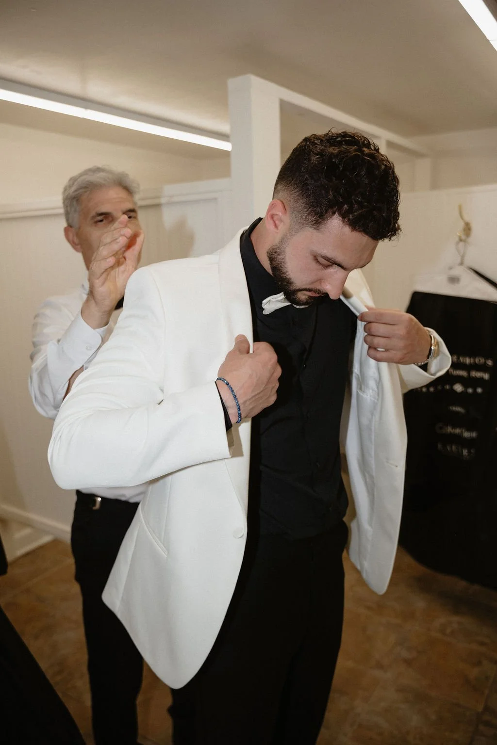 A man in a white tuxedo adjusting his jacket as another man in a white shirt stands behind him, holding his shoulder gently, in an indoor setting.