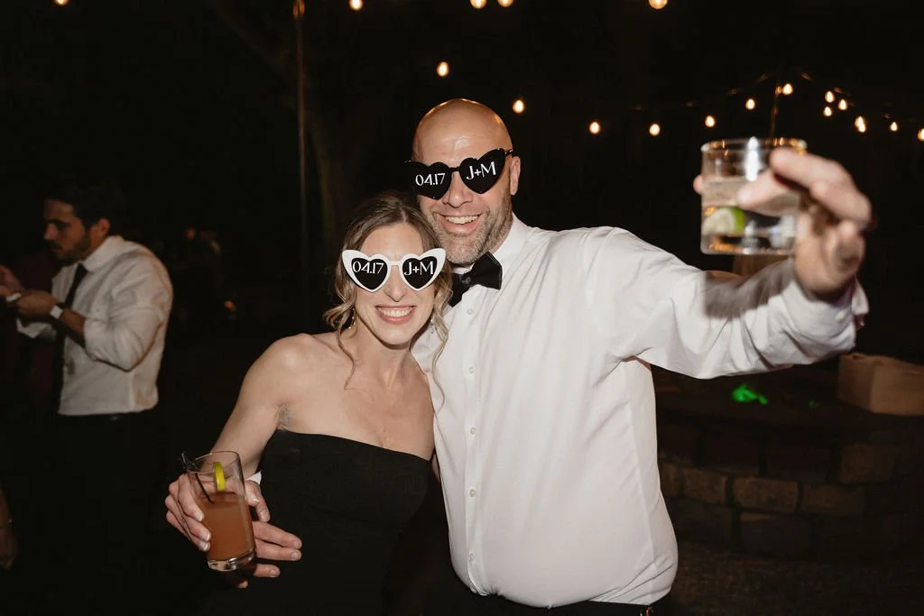 A man and woman dressed formally, wearing sunglasses with the initials "J+M" and the date "04.17" on them, smiling and holding drinks at a celebration or wedding reception.