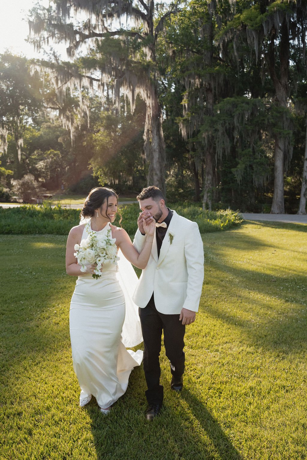 A bride and groom walking together on a grassy area outdoors, smiling and holding hands, with large trees and sunlight in the background.