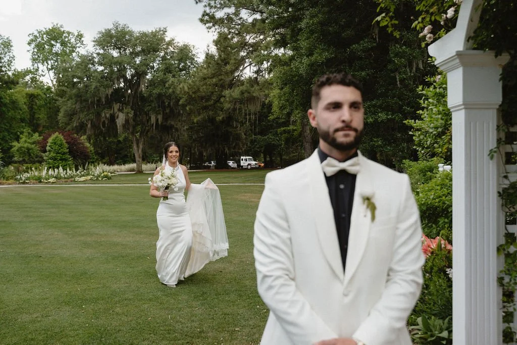 A bride holding a bouquet of flowers is walking towards a groom who is standing in front of a garden arch under an outdoor setting. The groom is dressed in a white tuxedo jacket with a black shirt and bow tie. The scene appears to be part of a weddin