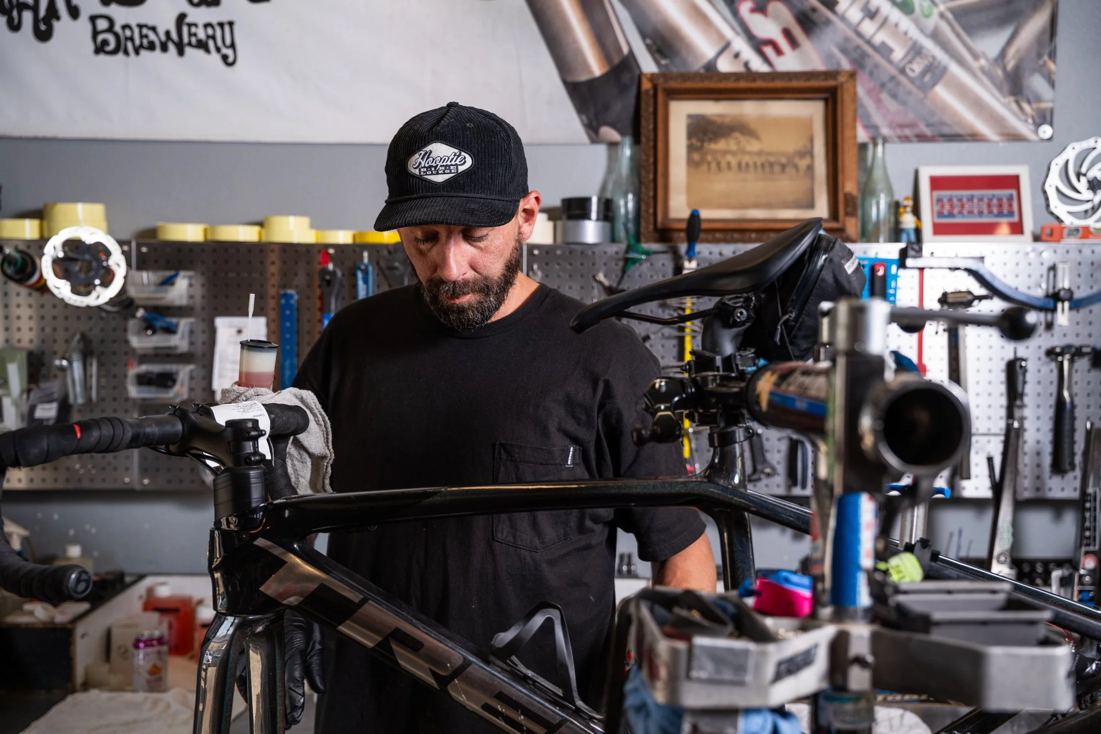 A man with a beard wearing a black cap and black shirt working on a black bicycle in a workshop, with tools and shelves in the background.