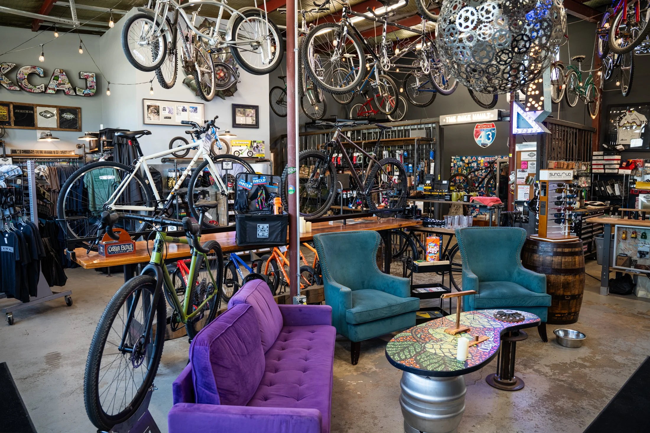 A store interior featuring numerous bicycles hanging from the ceiling, a purple velvet sofa, turquoise chairs around a colorful table, and various shelves with merchandise.