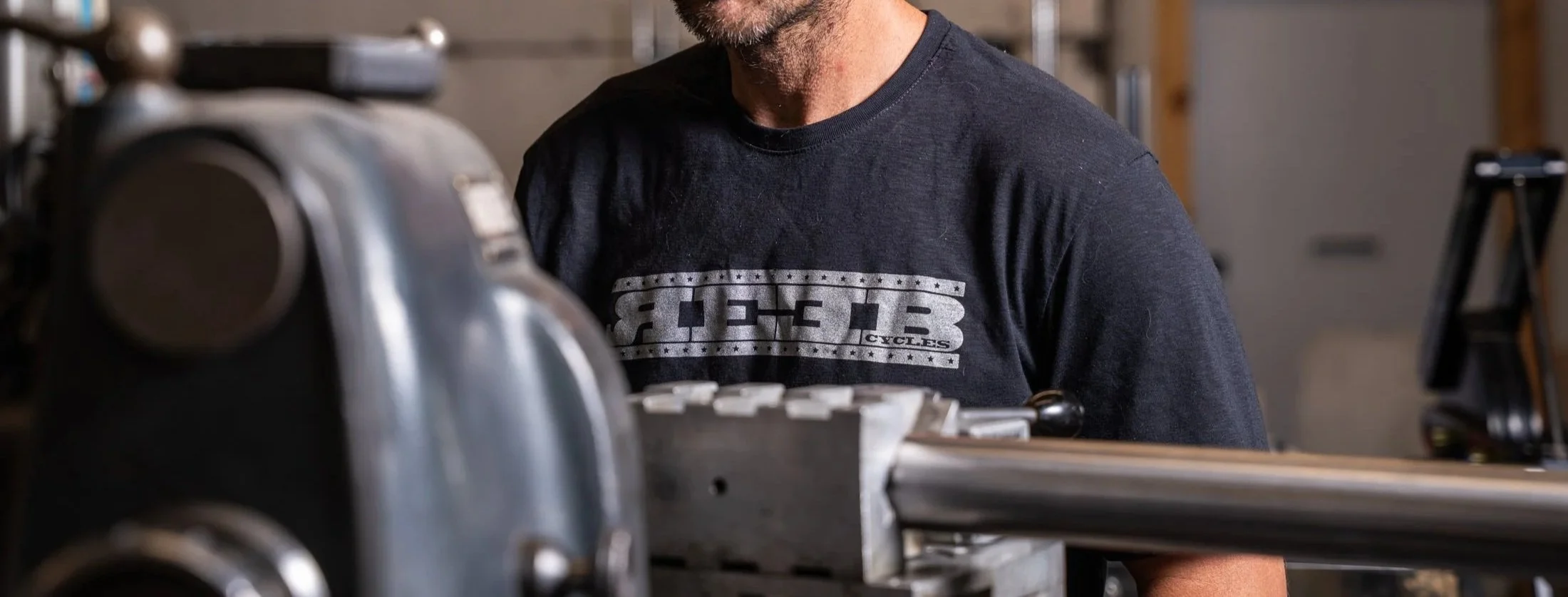 A man wearing a dark gray T-shirt with a graphic design, working on a piece of machinery in a workshop or garage.