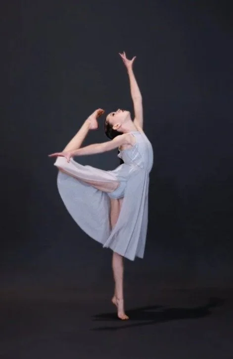 A ballet dancer in a light dress performing a dance pose against a black background.