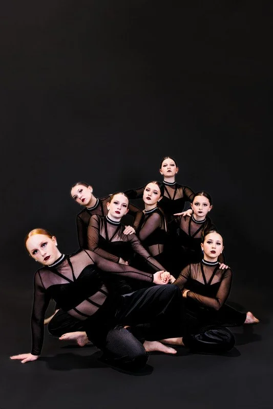 A group of six young female dancers in black sheer outfits with dark makeup poses on a black background.
