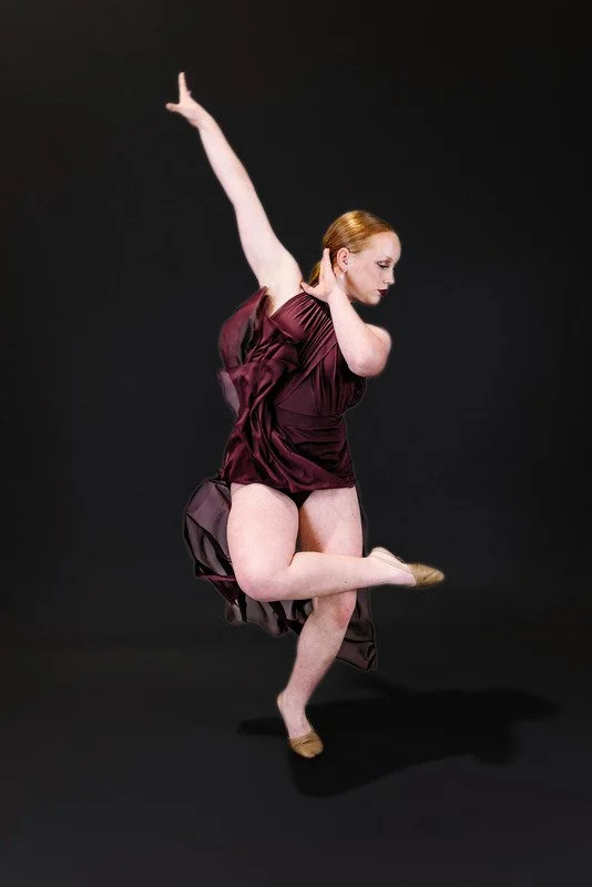 A woman with red hair in a maroon dress performing a dance move against a black background.