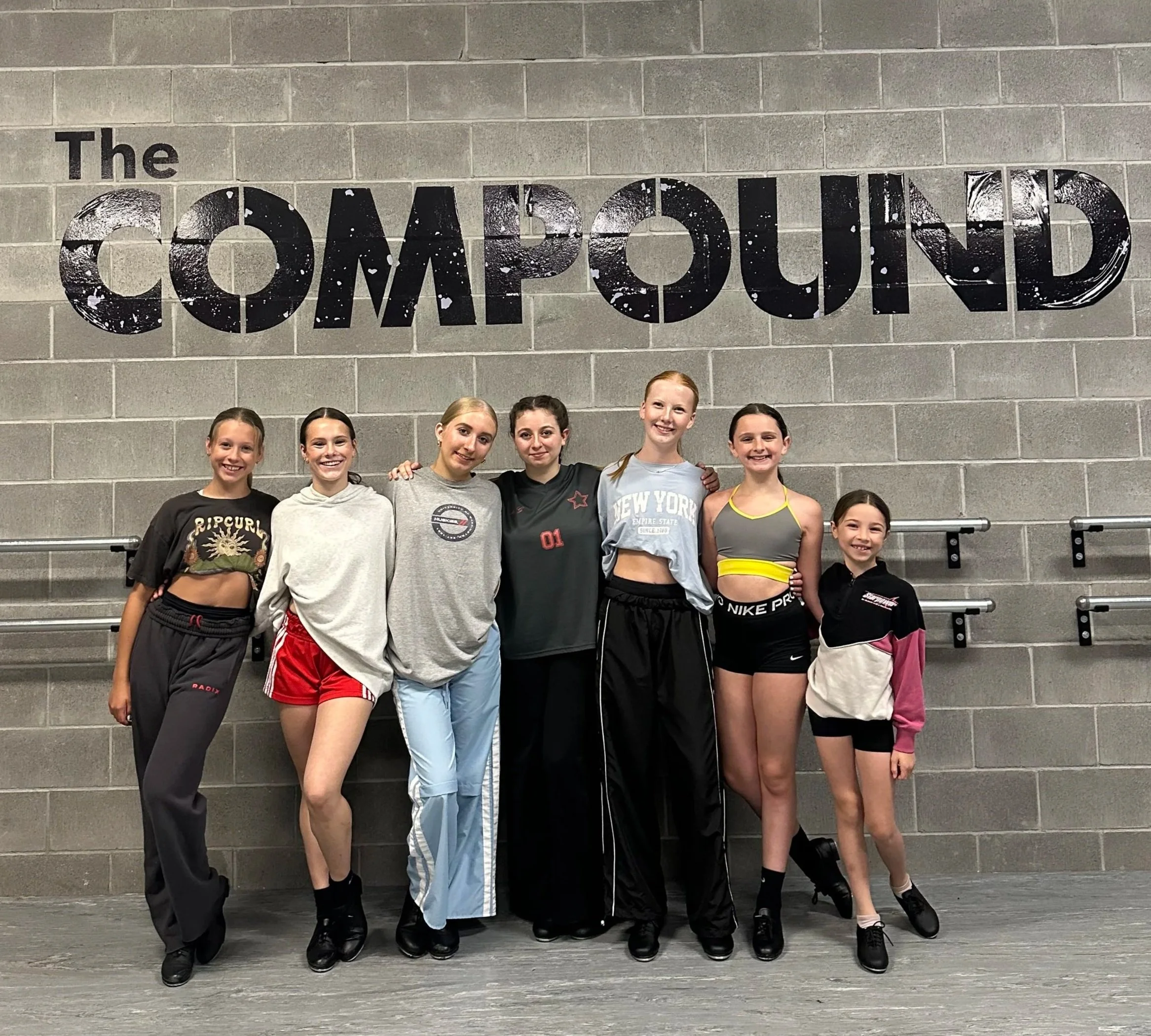 A group of six young girls smiling and standing close together in front of a grey brick wall with the words 'The COMPOUND' displayed in large black and white letters.