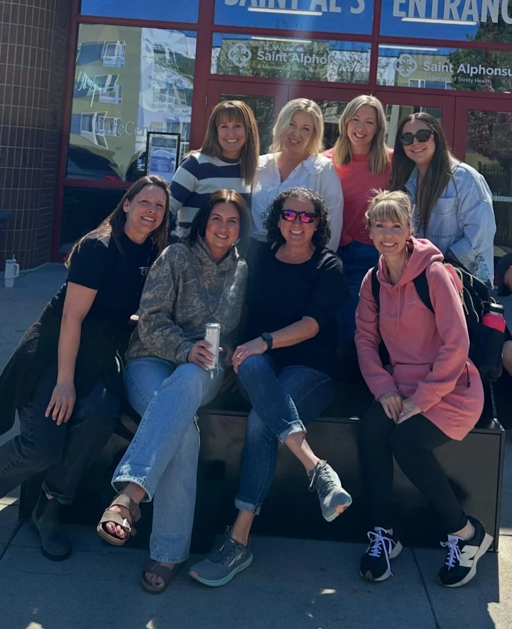 Group of nine women sitting and standing outside a building with red doors and glass windows, smiling for the camera.
