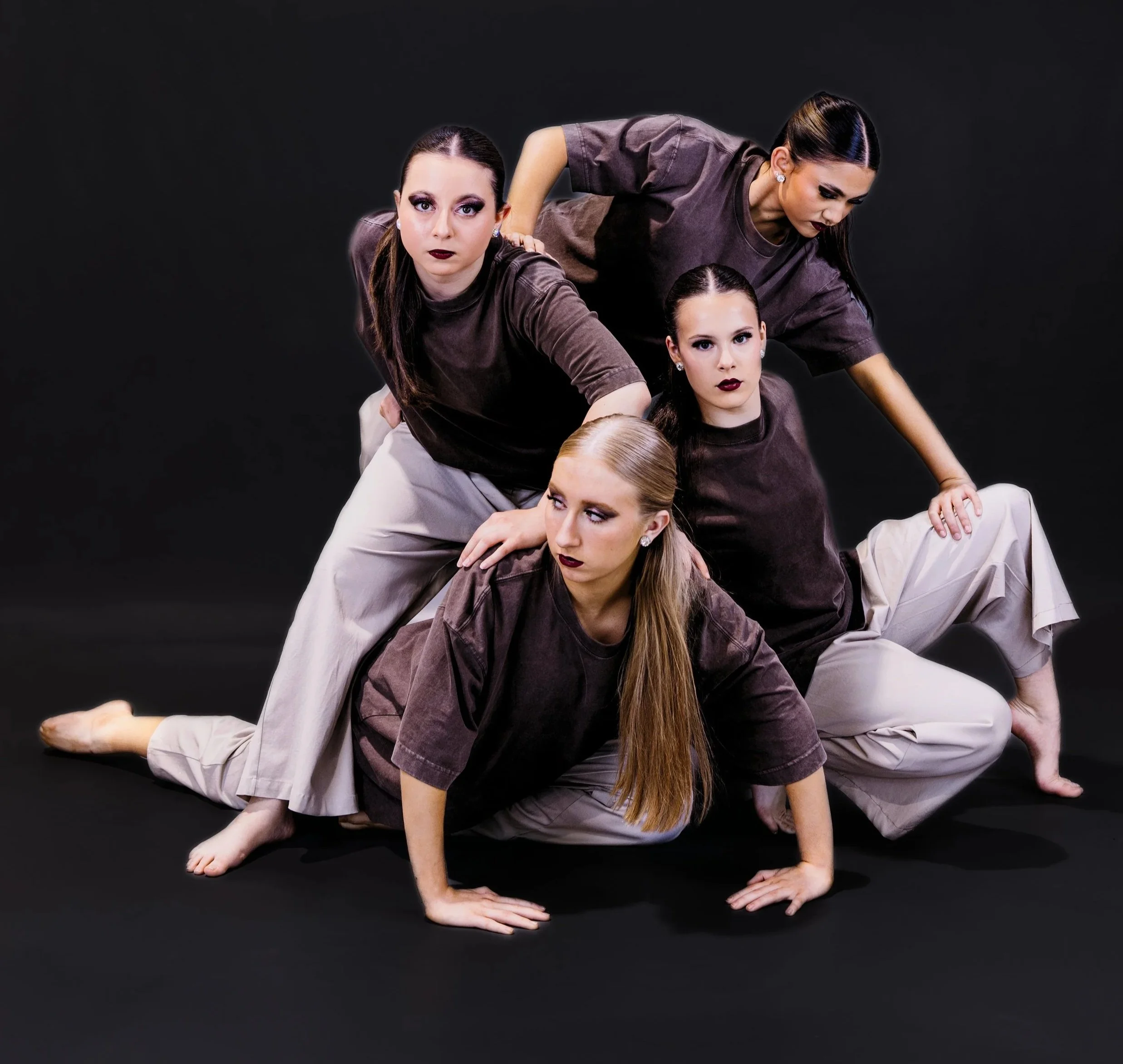 Four women dressed in gray tops and light-colored pants perform a complex yoga or acrobatic pose on a black background. Two women are supporting others in a layered formation, with focused expressions.