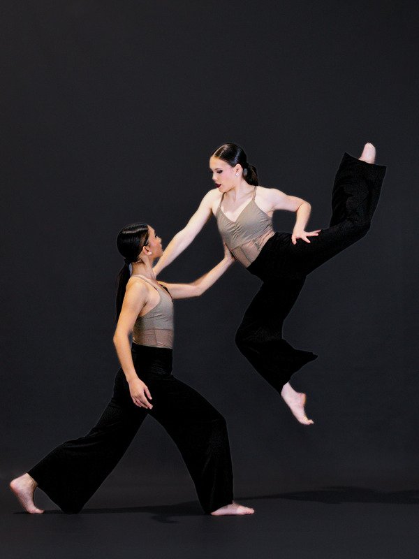 Two female dancers practicing dance moves against a black background. One is standing and the other is airborne in a leap.